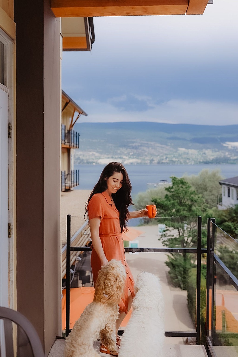 Guest holding a drink on her balcony while her dog stands beside her, with a scenic lake view.