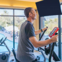 Man using an elliptical machine in a fitness room with large windows overlooking the resort pool.