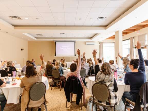 Attendees seated at round tables in a bright meeting room, raising their hands toward a presentation screen at the front.