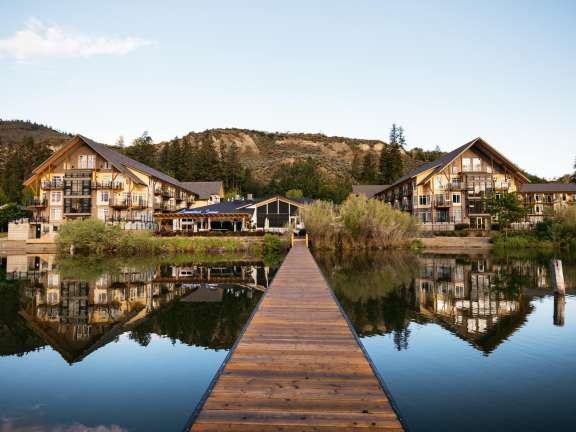 Wooden dock leading to Summerland Waterfront Resort buildings reflected in calm lake at sunrise