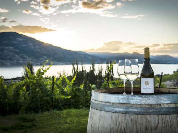 Wine bottle and glasses resting on a wooden barrel overlooking vineyards and Okanagan Lake.