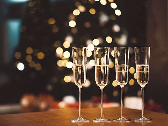 Four champagne flutes filled with sparkling wine on a wooden table, with blurred christmas tree & lights in the background.