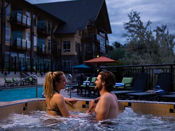 Couple enjoying an outdoor hot tub at dusk with lounge chairs and resort rooms behind them