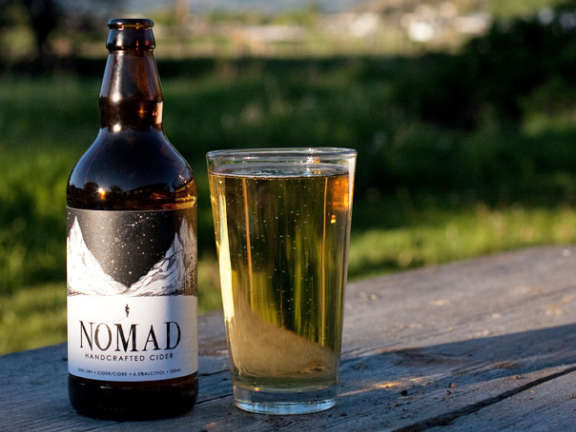 A bottle of Nomad Cider and a glass of cider on a wooden outdoor table with greenery behind it.