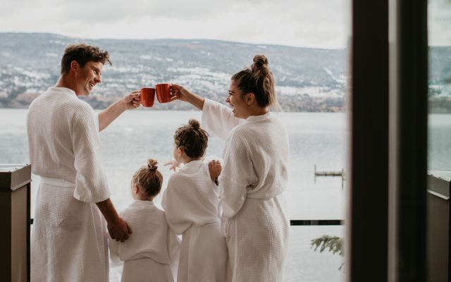Family of four with Mom, Dad, and two small daughters overlooking Okanagan Lake, enjoying coffee and hot chocolate in their robes on the balcony of their suite at Summerland Waterfront Resort.