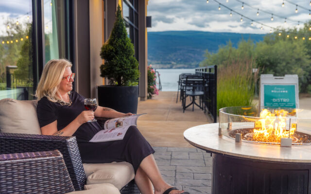 A woman in black dress seated on a cushioned patio chair at SWR, enjoying wine and reading near a fire table with lake views in the background.
