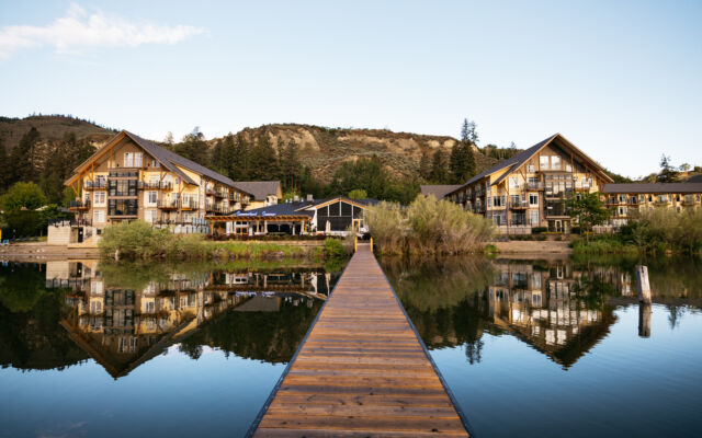 Wooden dock leading toward lakeside resort buildings reflected in calm water.