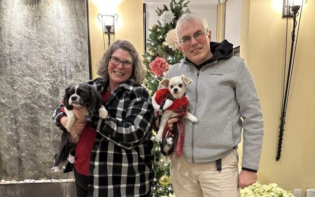 John and Kim of Hidden Gems of BC posing with their dogs, Mikki and Merlot, in front of a holiday tree and water feature in the Summerland Waterfront Resort lobby.