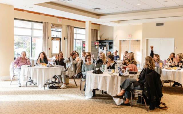 Retreat attendees smiling and applauding at round tables in the sunlit Waterfront Ballroom at Summerland Waterfront Resort & Spa in Summerland, BC